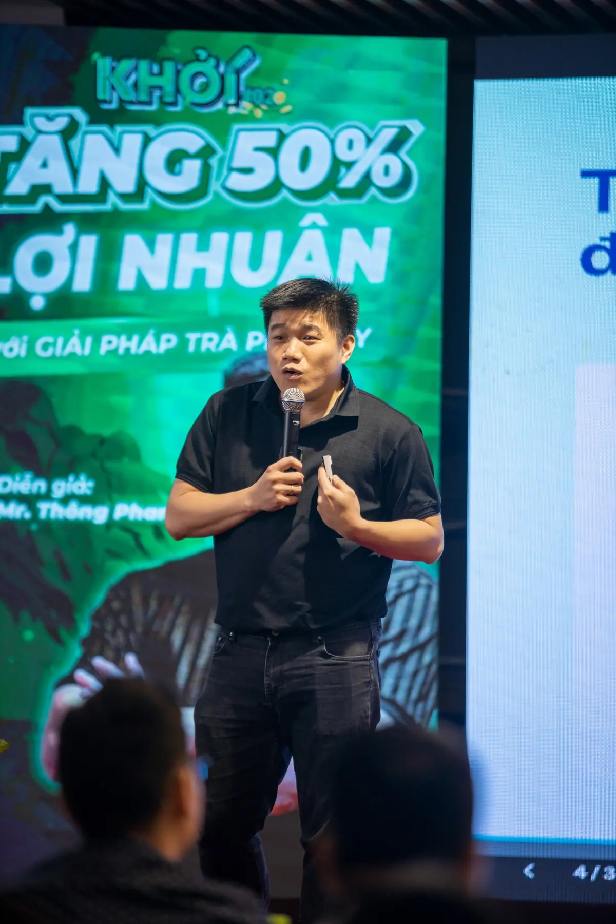 Chân dung điện ảnh của Thông Phan đang cầm micro chia sẻ trên sân khấu.